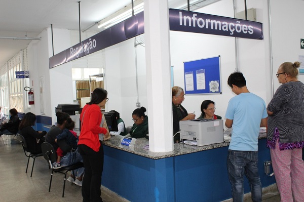 #PraCegoVer: Munícipes são atendidos na recepção da UBS, com funcionárias atrás da bancada, com placas acima dizendo 